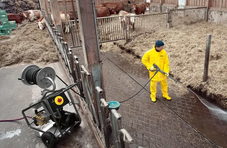 Farmař v ochranném obleku umývá podlahu stáje vysokotlakou čističkou.