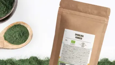 Balíček spiruliny vedle lžičky s práškem na bílém pozadí.