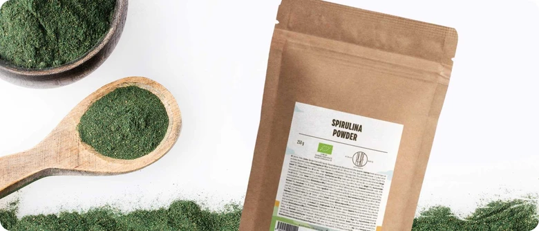 Balíček spiruliny vedle lžičky s práškem na bílém pozadí.